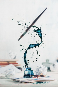 Spilled ink flying above inkwell in a spiraling splash with tiny drops and flying pen on a light background. Still life with writer workplace. Creative writing concept. Dynamic shot with frozen motion.
