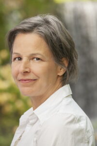 Cheryl Wilder is a middle-aged woman with a mix of gray and brown hair cut short, wearing a white-collared shirt, standing in front of a waterfall and green foliage with her head turned to the left and looking into the camera