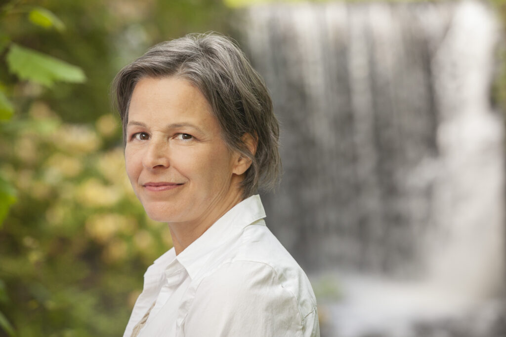 Cheryl Wilder is a middle-aged woman with a mix of gray and brown hair cut short, wearing a white-collared shirt, standing in front of a waterfall and green foliage with her head turned to the left and looking into the camera