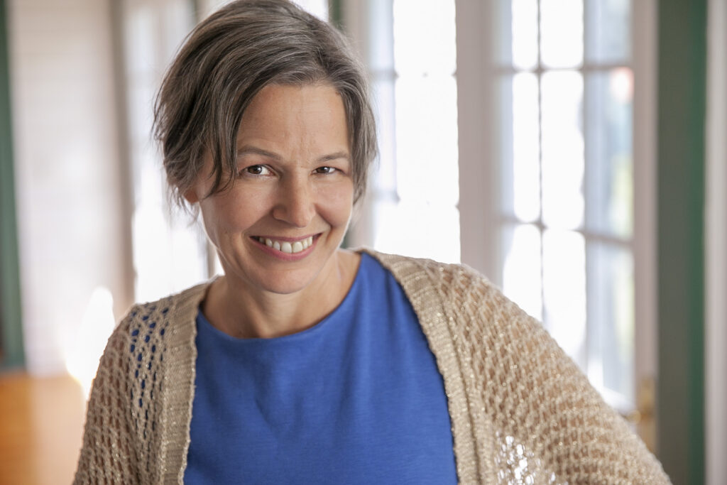 Cheryl Wilder is a middle-aged woman with a mix of gray and brown hair cut short, wearing a royal blue dress and a shimmery gold woven cardigan standing in front of a row of windows