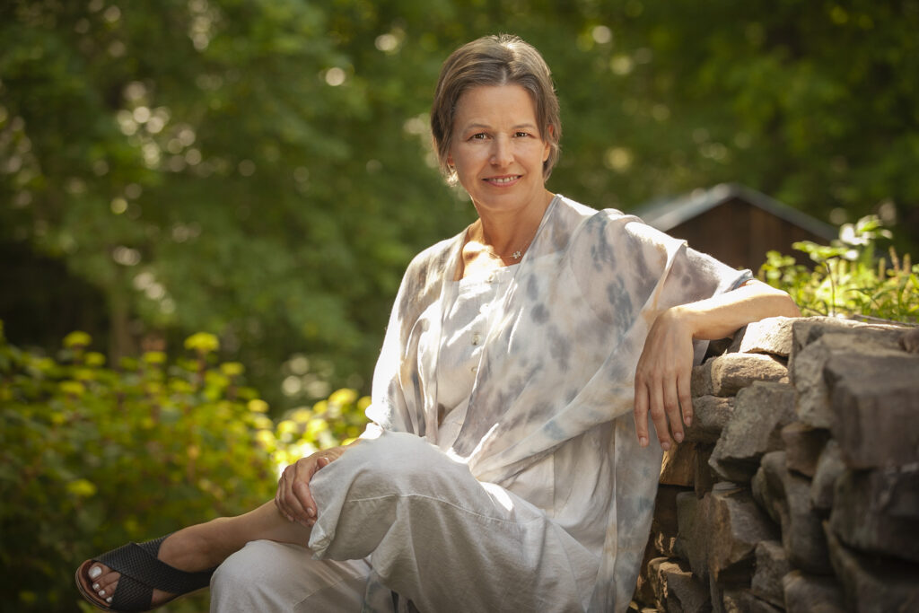 Cheryl Wilder is a middle-aged woman with a mix of gray and brown hair cut short, wearing a white jumpsuit and a blue, white, and tan light wrap sitting by a rock wall in a tree-laden natural setting