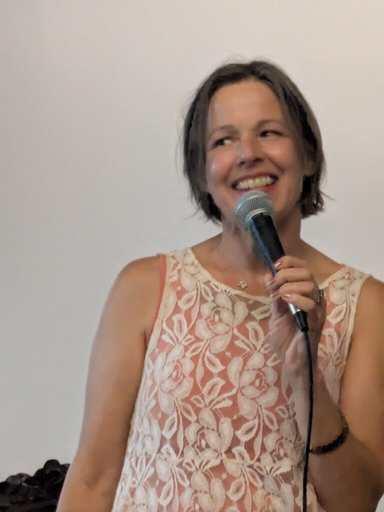 White woman with chin-length brown hair wearing a lace sleeveless slip over a rust colored skirt holding a microphone and talking