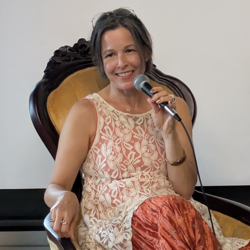 White woman with chin-length brown hair sitting in an antique high back chair with with mustard yellow fabric wearing a lace sleeveless slip over a rust colored skirt holding a microphone and talking